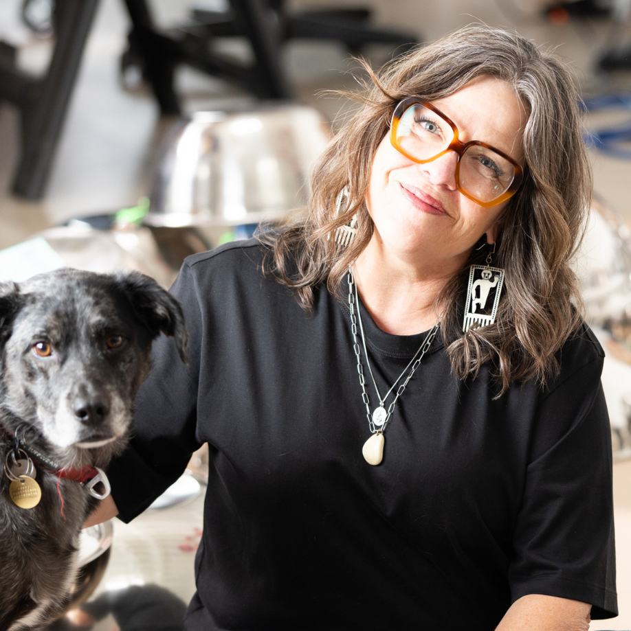 An image of Marie Watt in her studio. She is sitting on the floor petting her dog. Behind her is her studio with several art supplies sitting around the space. Marie is an olive medium complexion. She is wearing glasses, long silver dangle earrings, several silver necklaces, and several turquoise bracelets on her left arm. She is wearing a black t-shirt and blue jeans. She is sitting cross-legged. The dog is dark with short shaggy hair. The dog has red collar with a gold medallion.
