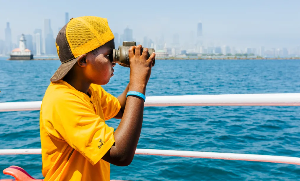 A young boy with a deep skin tone stands on a boat looking through binoculars. He wears a bright yellow t-shirt and a backwards yellow hat. Behind him there is blue water and the Chicago city skyline. 