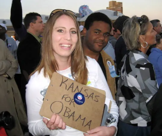 Clarissa Unger, a woman with a light skin tone, faces front while smiling at the camera; her hair is a golden dark blonde and she has a pair of black sunglasses on her head. She is wearing a white long-sleeved t-shirt, in her hand she is carrying a clipboard and a small sign that says, “Kansas for Obama behind her is a brown-skinned man also smiling into the picture, he is wearing a beige suede jacket with a black shirt underneath, on his lapel is an Obama for Kansas button. The overall background of the picture is an outdoor rally.