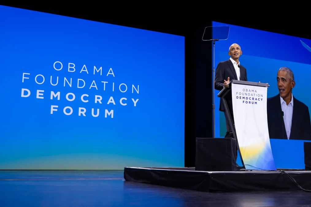 President Obama stands behind a podium at the Obama Foundation Democracy Forum. The front of the podium reads, “Obama Foundation Democracy Forum.” A screen in the background says the same.