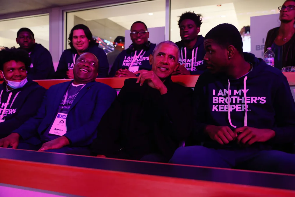 Two rows of seated people, purple arena lights illuminate President Obama and young boys in sweatshirt that say"I AM MY BROTHER'S KEEPER' during a Chicago Bull's basketball game.