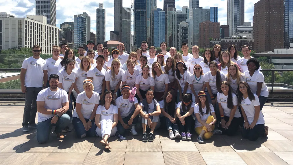 A group of young people all wearing white t-shirts with the same logo sit and stand in a semicircle, posing for the camera. Behind them is a city skyline. It is a sunny day and shadows are cast on and around the group.