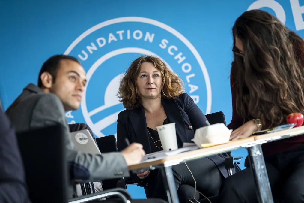 Three people sit around a table. A woman in the center with light skin and wavy light brown hair looks to the right. Behind them is an Obama Foundation Scholars logo. 