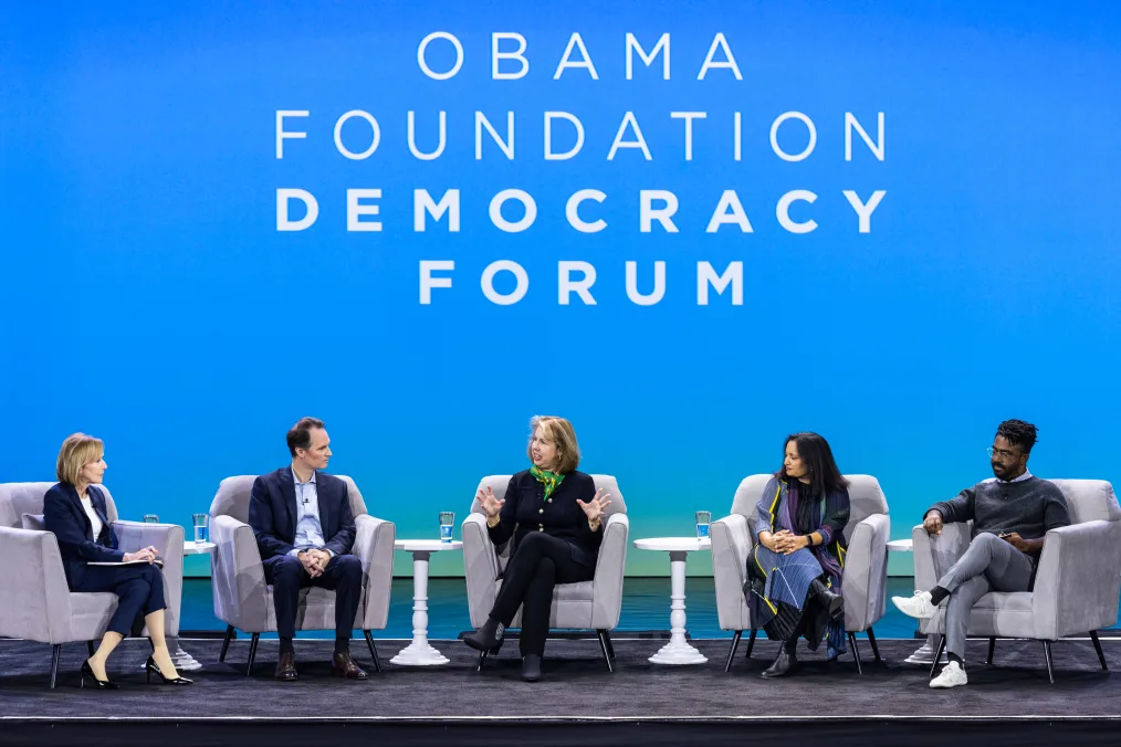 Nancy Gibbs, a woman with a light skin tone, sits center stage alongside John Palfrey, a man with a light skin tone,  Sonal Shah, a woman with a medium skin tone, Darryl Holliday, a Black man with a deep skin tone and locs, and Judy Woodruff, a woman with a light skin tone and short brown hair. The screen behind them reads, “Obama Foundation Democracy Forum.”