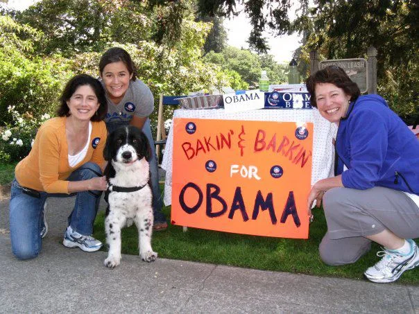 Three light-skinned, dark-haired women crouch down by their black-and-white  dog and an orange sign that reads "Bakin' and Barkin' for OBAMA."