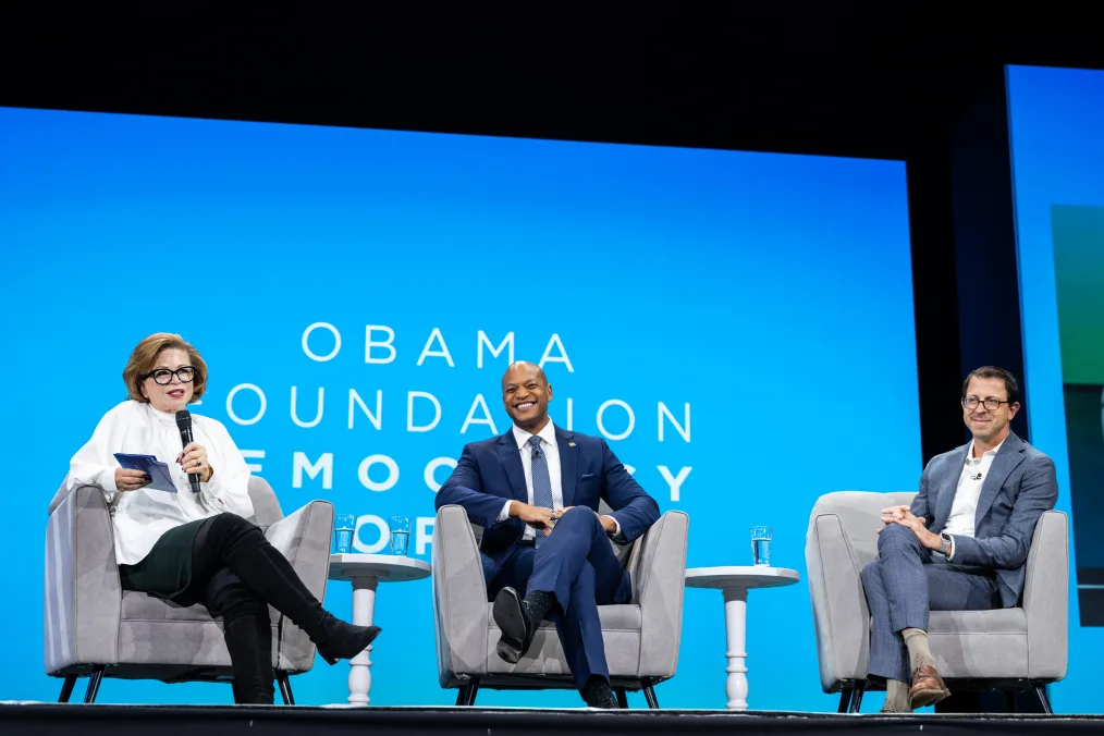 Valerie Jarrett sits on a panel alongside Maryland Governor Wes Moore, a Black man with a medium skin tone and no hair, and Ian Bassin, a man with a light skin tone who wears glasses. The screen behind them reads, “Obama Foundation Democracy Forum” in white text against a cyan background.