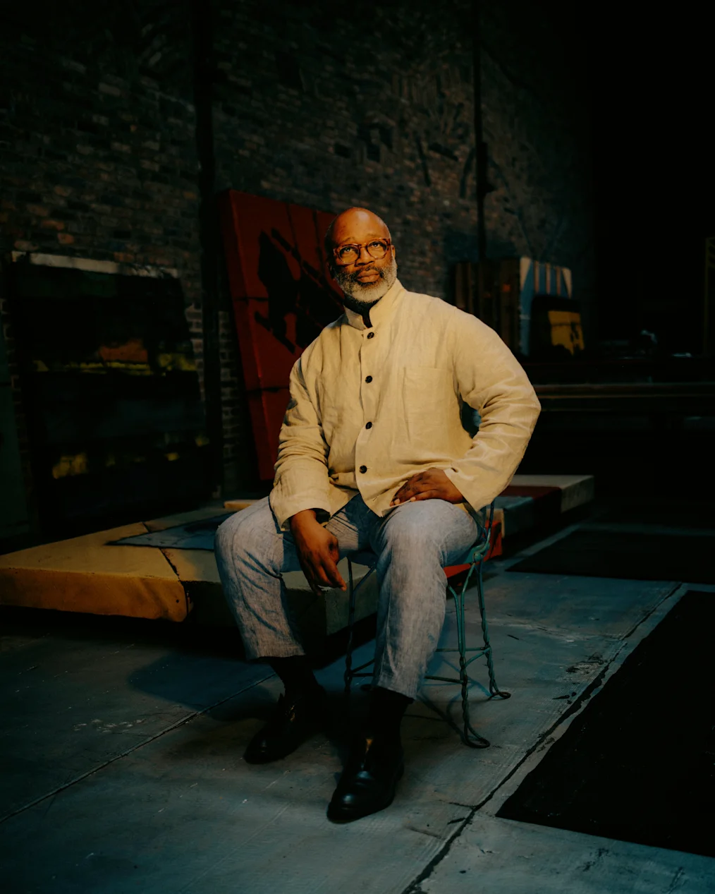 Photograph of Theaster Gates in his art studio. The background is dark. There is a brick wall to the right of Theaster. In front of the wall is a work cabinet and a large red installation. Theaster is sitting on a large wooden work table. Theaster has a medium deep skin tone and a no hair. He has a gray, salt and pepper beard. He is wearing red rectangular glasses. His shirt is light colored, long sleeved. He is wearing blue jeans and dark shoes.​​​​‌﻿‍﻿​‍​‍‌‍﻿﻿‌﻿​‍‌‍‍‌‌‍‌﻿‌‍‍‌‌‍﻿‍​‍​‍​﻿‍‍​‍​‍‌‍​﻿‌‍﻿﻿‌‍﻿‍‌﻿‌​‌‍‌‌‌‍﻿‍‌﻿‌​‌‍‌‍‌﻿‌‌‌‍﻿​​‍﻿‍‌‍​﻿‌‍﻿﻿‌‍﻿‌​‍​‍​‍﻿​​‍​‍‌‍‍​‌﻿​‍‌‍‌‌‌‍‌‍​‍​‍​﻿‍‍​‍​‍‌‍‍​‌﻿‌​‌﻿‌​‌﻿​​‌﻿​﻿​﻿‍‍​‍﻿﻿​‍﻿﻿‌‍​‌‌﻿​​‌﻿​​​‍﻿‍‌‍​﻿‌‍﻿﻿‌‍﻿‍‌﻿‌​‌‍‌‌‌‍﻿‍‌﻿‌​‌‍‌‍‌﻿‌‌‌‍﻿​​‍﻿‍‌‍​﻿‌‍﻿﻿‌‍﻿‌​‍﻿﻿‌﻿​﻿‌﻿​​‌‍​‌‌‍​﻿‌‍‌‌‌﻿​﻿​‍﻿﻿‌‍﻿​​﻿‌﻿‌‍‍​​﻿‌‌​﻿‍‌‌‍‍​‌‍‌‍‌‍﻿‍‌‍﻿​‌﻿‍​‌‍‍‍‌﻿‍​​‍﻿﻿‌‍‌‌‌‍﻿‍‌﻿‌‍‌‍‍‌‌﻿​‍‌‍﻿﻿‌‍﻿‍‌‍﻿‌‌‍‌‌‌‍﻿‍‌﻿‌​‌﻿​﻿​‍﻿﻿‌‍﻿‌‌‍​‌‌﻿​﻿‌﻿‌​‌‍‌‌‌﻿​‍​‍﻿﻿‌‍‌‌‌‍﻿‍‌﻿‌​‌﻿​‍‌‍‍‌‌‍‌‌‌﻿​﻿​‍﻿﻿​﻿‌​‌​﻿‌​﻿​﻿‌​‍‍​﻿‌‍‌‌​﻿​﻿‍‌‌‌‍‍‌‌​﻿‌‌​﻿‌‍‌​‌​‍﻿‌​﻿‌‌﻿​​‌﻿‌‌‌​‌﻿‌﻿‍​‌‌​‌‌‍​‌‌﻿​‌‌​‌‌‌‍‌﻿​‍﻿﻿​﻿﻿﻿‌‍‌‍‌‍﻿﻿‌‍​﻿‌﻿‌‌‌﻿​﻿‌‍‌‌‌‍‌​‌​‌‍‌‍‍‌‌‍‌‌‌‍﻿​‌‍‌​​﻿﻿‌‌‍​‌‌‍﻿​‌﻿‌​‌‌‌​‌‍‌‌‌﻿‍​‌﻿‌​​‍‌‍‌‍‌‍‌‍﻿﻿‌‍​﻿‌﻿‌‌‌﻿​﻿‌‍‌‌‌‍‌​‌​﻿​‌‍﻿﻿‌‍​﻿‌‍​‌‌‍﻿​‌‍‌‌​﻿﻿‌‌‍‌‌‌‍﻿‍​‍﻿‌‌‌‌‌‌‌​﻿​‍‌‍‌﻿​﻿‌‍﻿﻿‌﻿‌‌‌﻿​‍‌‍​﻿‌‍‌‌​﻿﻿‌‌﻿‌‍‌‍‌‌‌﻿​‍‌‍​﻿‌‍‌‌‌‍﻿​​‍﻿‌‌‍​﻿‌‍﻿﻿‌‍﻿‍‌﻿‌​‌‍‌‌‌‍﻿‍‌﻿‌​​‍﻿‌‌‍﻿​‌‍‍‌‌‍﻿‍‌‍‍﻿​‍​‍​‍﻿​​‍​‍‌‍​﻿‌‍﻿﻿‌‍﻿‍‌﻿‌​‌‍‌‌‌‍﻿‍‌﻿‌​‌‍‌‍‌﻿‌‌‌‍﻿​​‍​‍​﻿‍‍‌﻿‍﻿​‍​‍‌‍‌‌‌‍‌​‌‍‍‌‌﻿‌​‌‍﻿﻿‌﻿​‍‌​‍‌‌‍﻿‍‌﻿‌​‌‍‌‌‌﻿​‍‌‍‌‍‌‍​‌‌‍​﻿‌‍‌‌​‍​‍​﻿‍‍‌﻿‍﻿​‍​‍‌﻿‌﻿‌‍‍‌‌‍‌​‌‍‌﻿‌‍‌‌‌﻿‌​‌​‍‌‌‍‌​​‍​‍​﻿‍‍​‍​‍‌‍﻿‌‌﻿‌‌‌‍﻿​‌﻿‌​‌‍‍‌‌﻿​​‌‍﻿​‌‍‌‌‌​﻿​‌‍‍‌‌‍﻿‍‌‍‌‌​‍​‍​‍﻿​​‍​‍‌﻿‌﻿‌‍‍‌‌‍‌​‌‍‌﻿‌‍‌‌‌﻿‌​‌​﻿‍‌‍​‌‌‍﻿‌‌‍‌‌‌﻿​﻿‌﻿​​‌‍​‌‌‍​﻿‌‍‌‌​‍​‍​﻿‍‍​‍​‍‌‍​‍‌﻿‌‌‌‍‍‌‌‍﻿​‌﻿‌​‌‍‍‌‌‍﻿‍​‍​‍‌﻿﻿‌​‍﻿​​‍​‍‌‍‌‍‌‍‍‌‌‍‌‌‌‍﻿​‌‍‌​‌‌‌​‌﻿‍‌‌﻿​​‌‍‌‌​‍​‍​﻿‍‍​‍​‍‌‌‌​‌‍‌‌‌﻿‍​‌﻿‌​​‍​‍‌﻿﻿‌‌﻿﻿‌
