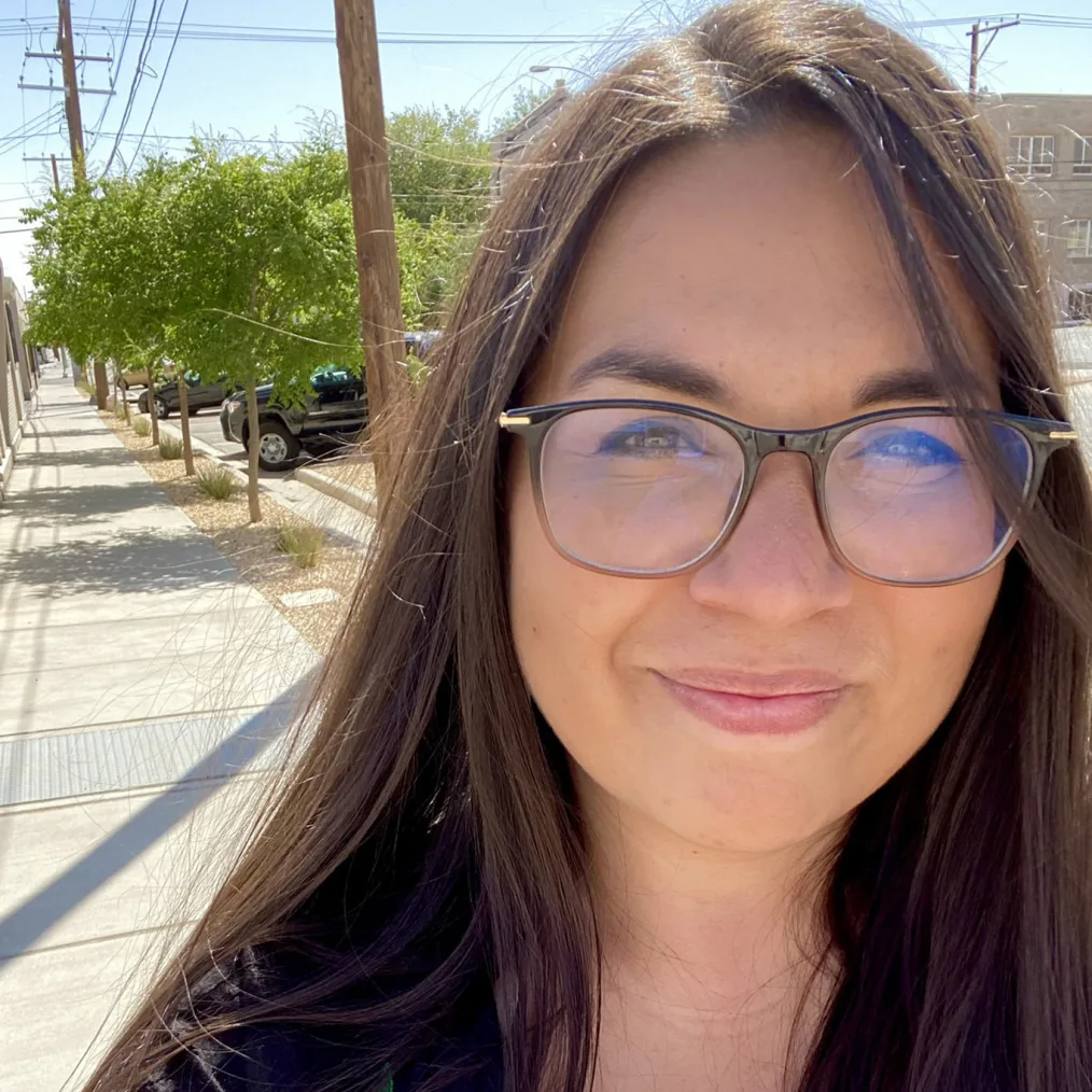 ethany Rivera Molinar, a Latina woman with a medium skin tone, holds a closed-lipped smile. She stands outdoors on a sunny day. She has long brown hair and clear glasses. 