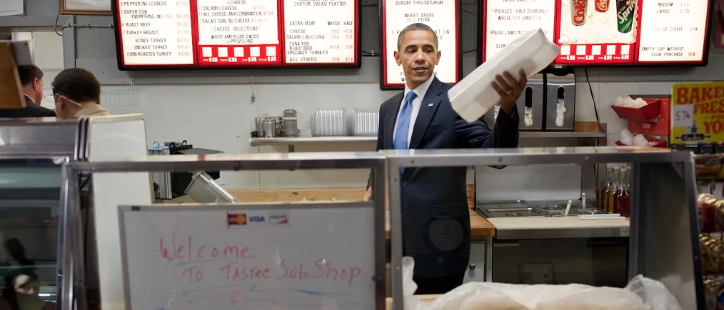 President Barack Obama, wearing a dark blue suit and tie, stands behind a counter holding a long white paper bag. There is a glass window in front of him with a sign that says "Welcome to Tastee SubShop". Behind President Obama are big screens displaying the menu for the sub shop with red borders. Two people are in the background on the left-hand side of the photo.