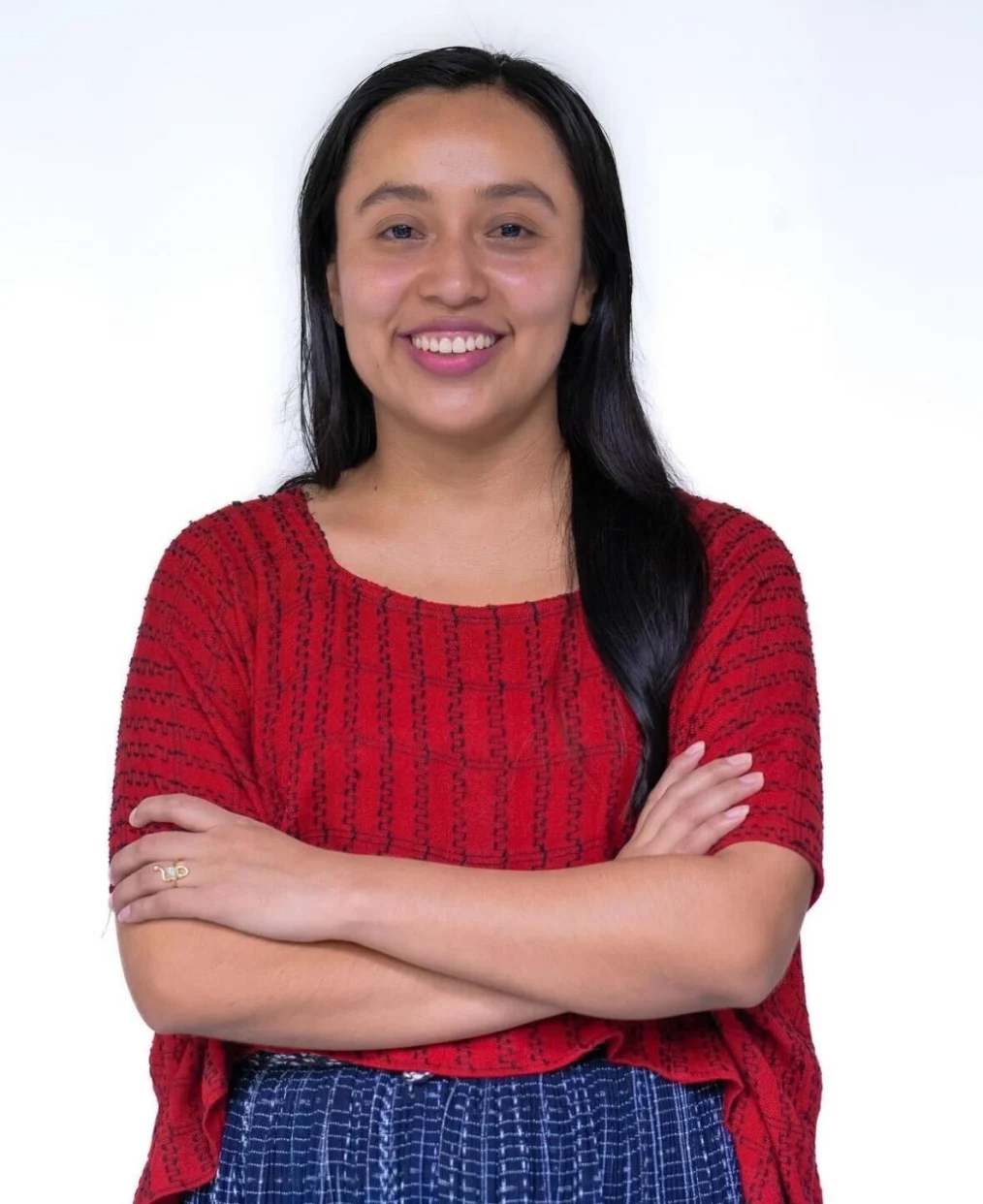 The image is a “cowboy shot” of a woman with a light skin tone and long black hair. She smiles at the camera. Her arms are crossed around her stomach. She is wearing a red indigenous print shirt with black dots and a blue patterned skirt. 