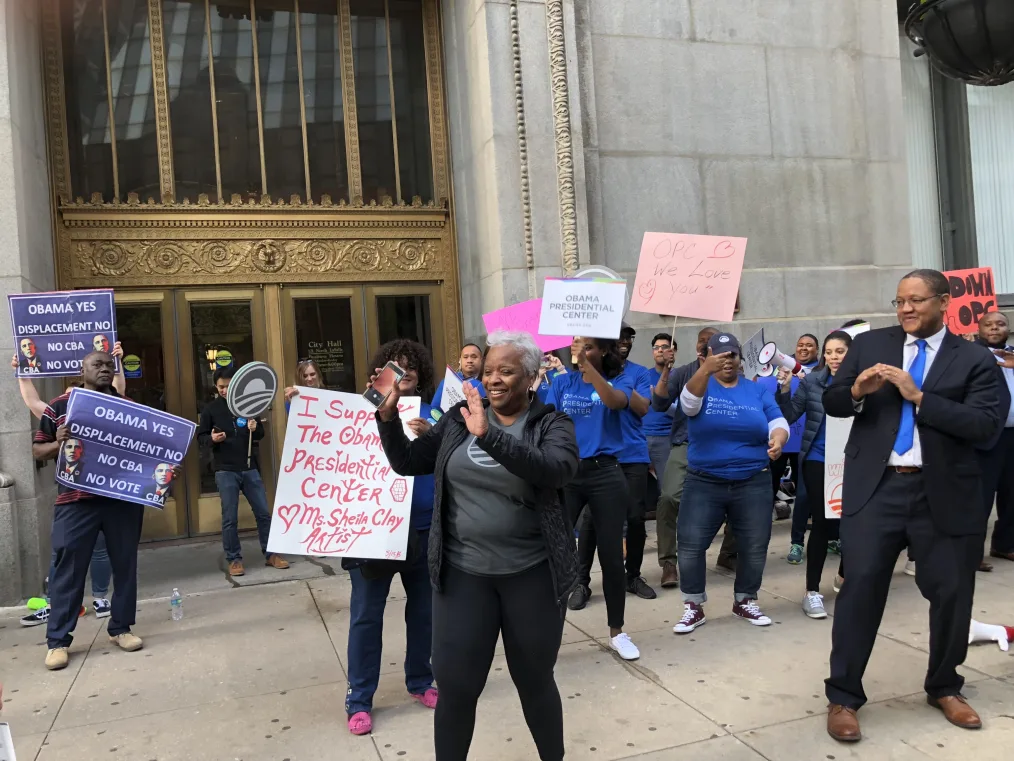 A group of people wearing blue Obama Presidential Center t-shirts appear to be dancing on a sidewalk as they hold signs supporting the Center. One woman wearing a gray Obama Foundation logo shirt stands in the center. To the left are men holding signs that read "Obama Yes, Displacement No, No CBA, No Vote."