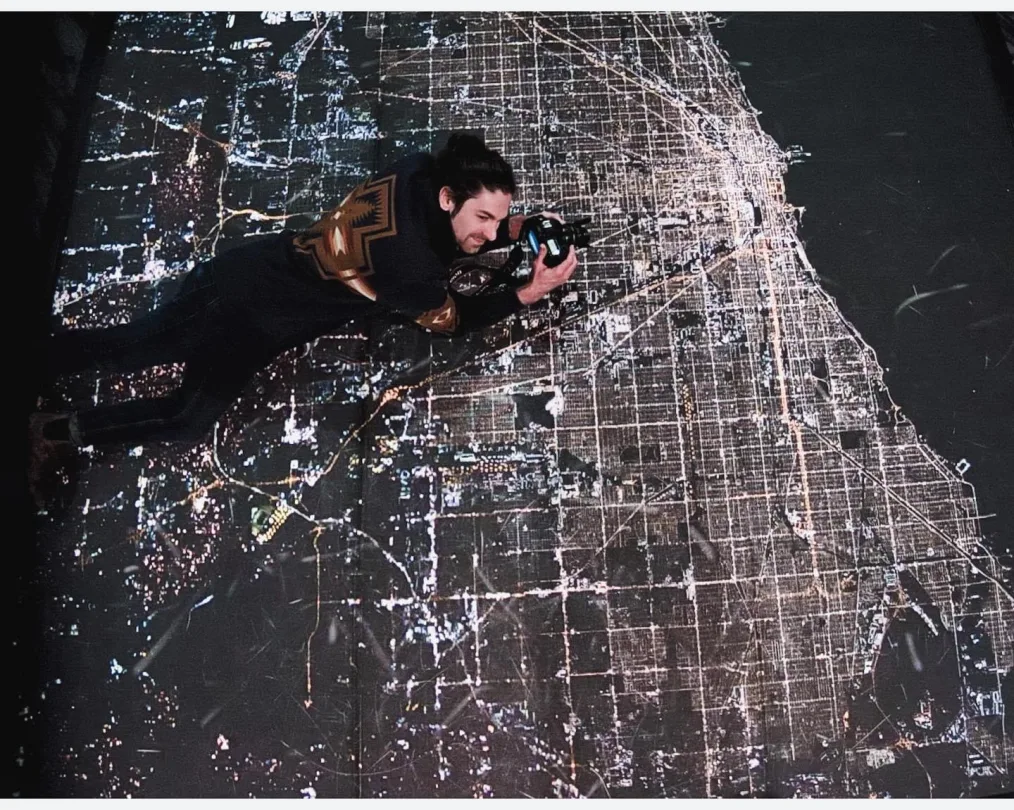 The image is a photo of Gregg Ross laying on a geographical map that is has varying stages of light on it. These various stages of light are meant to symbolize population density. Gregg has pale skin, a dark colored goatee, and long dark hair that is pulled into a bun. He is wearing a black shirt with a brown design on it and black pants. He is holding a camera.