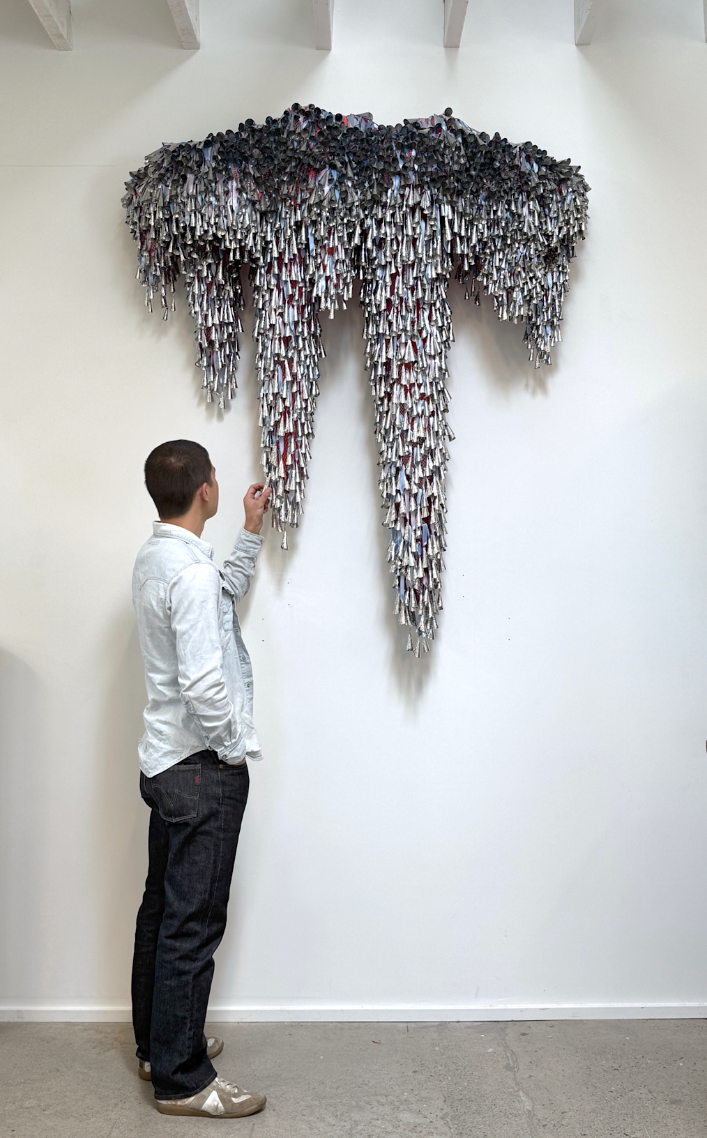 An image of a person looking at Marie Watt's installation Sky Dances Light. The person is facing away from the camera. They have closely cropped dark hair. They are wearing a light blue long sleeved shirt, dark pants and sneakers. The installation is made up of tin jingles attached to a white wall. The jingles are silver cones. There are thousands of them. In between the jingles are straps of pink and blue fabric.