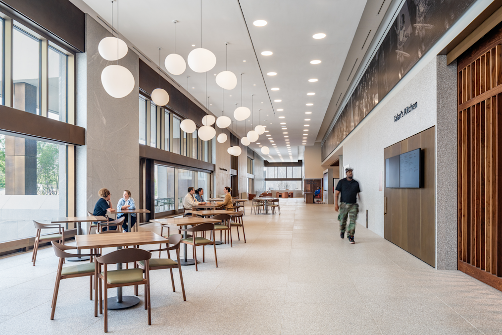 A horizontal photograph captures a wide, sunlit atrium corridor featuring polished light-colored flooring and large floor-to-ceiling windows along the left side. Hanging from the white ceiling, a cluster of glowing white, oval-shaped pendant lights extends down the center of the hallway. Next to them is a wall labeled "Tafari's Kitchen." 

