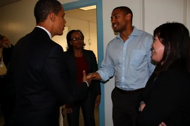 President Obama is shown shaking the hand of a young man with a deep skin tone while surrounded 
by other women with deep and medium skin tones.