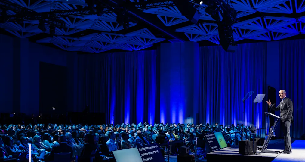 A large audience in a dimly blue lit room listens to President Obama Speak at the 2024 Democracy Forum. 