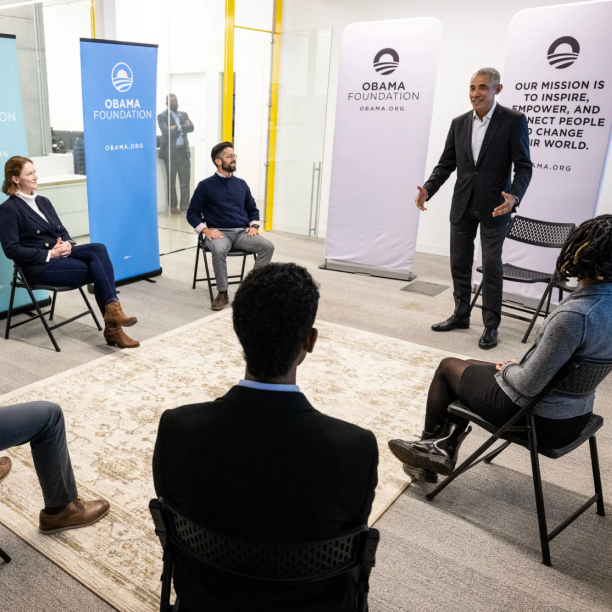 A photo of President Obama and a circle group of middle aged men and women. They are in a small office space, that has see in glass on the left. On the floor is a yellow/beige carpet with brown designs. Each person sits in a light black chair except President Obama who is standing and saying something. On their right are a set of spread out Obama Foundation Posters. Two men and women sit with light skin are sitting in the left of the photo in formal attire. On the right is one man and woman with medium to deep skin tones in formal attire. There faces are not visible to the camera.