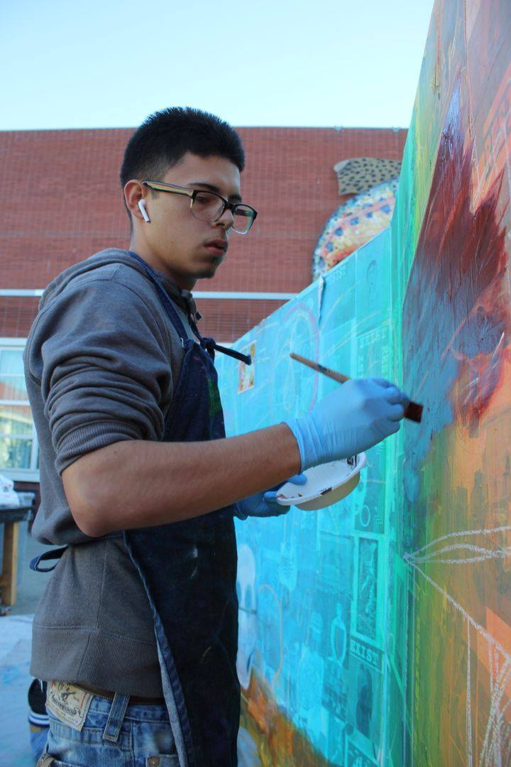 A young man with medium skin tone and glasses wears earbuds and an apron as he paints a colorful wall in front of him.