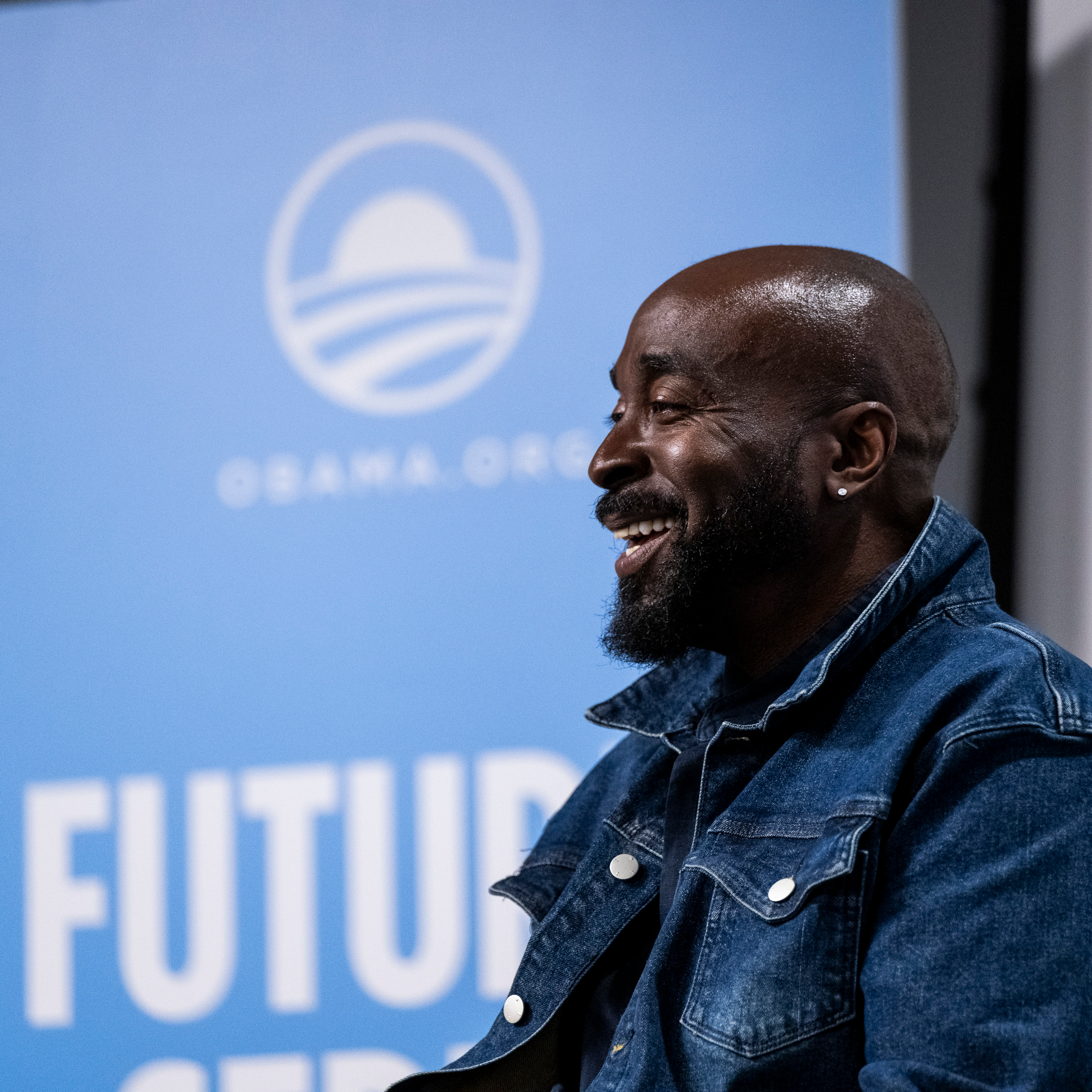 A side profile of Eric Williams, a Black man with a dark skin tone and no hair, smiling at an audience. He is wearing a denim shirt with a flipped collar. A blurred blue banner in the background reads, “Obama.org.”