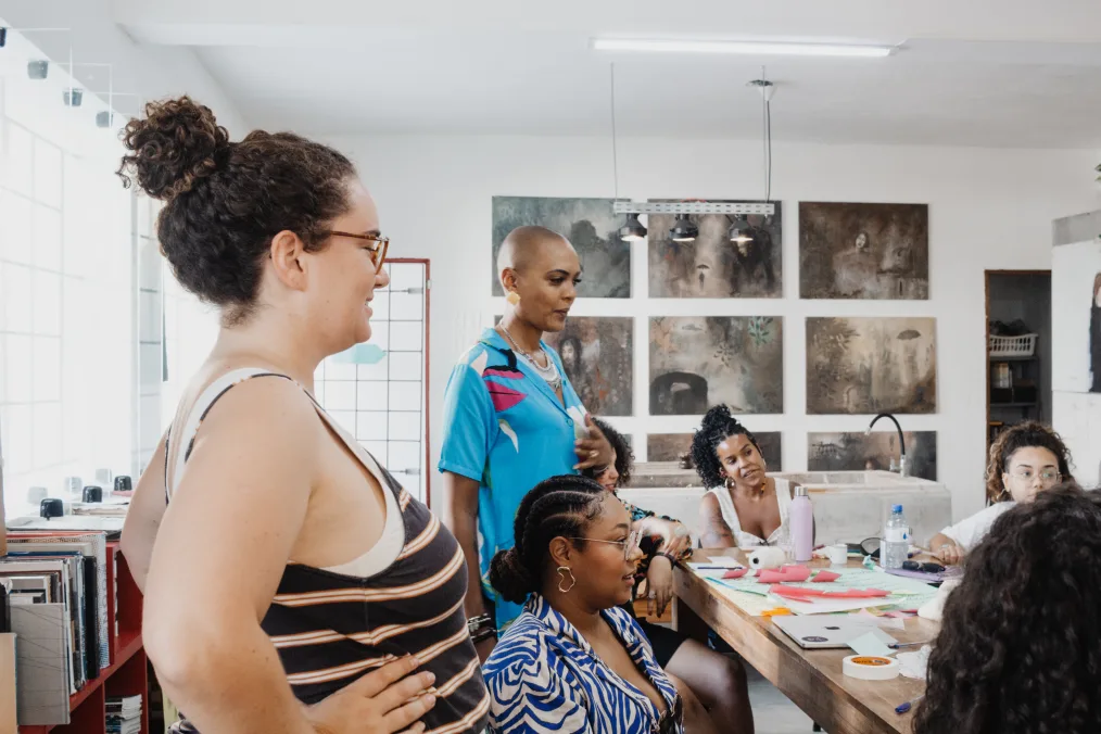 In a room, a woman with a light skin tone and curly hair rests her hands on her hips. She is surrounded by six other women with a range of ages and light to dark skin tones. There are poster boards and pink sticky notes on the table. All are looking away from the camera. 