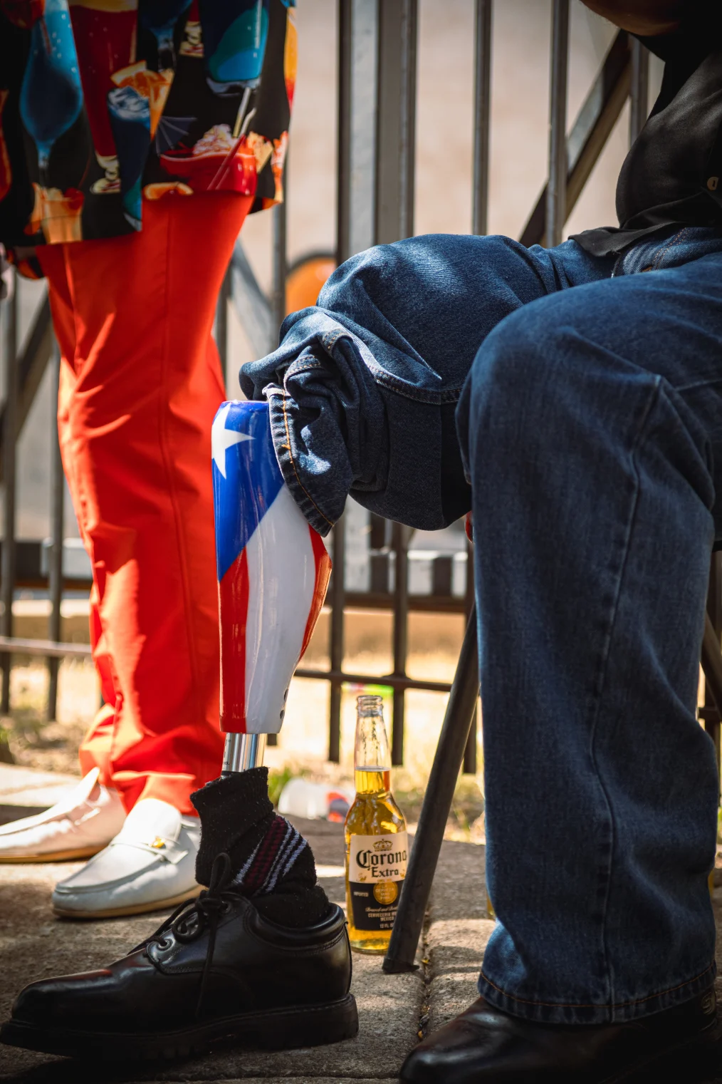 The image is of the feet of two people in front of a steel fence. The first person has on orange pants and white loafers. The second person is sitting on a chair: he has blue jeans on. One of the legs of his jeans is pulled up, revealing a prosthetic leg that is design with the Puerto Rican flag. On the ground between the two people is a Corona beer.