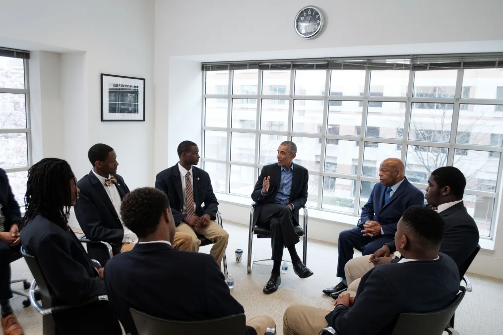 Barack Obama, dressed in a dark suit and blue dress shirt, sits in a chair alongside six young Black men and John Lewis who are also seated. In the background there is a clock above Barack Obama's head, a framed photo on the wall to the left and a large picture window behind him. The young men are dressed in dark suit jackets, white collared shirts and brown pants.