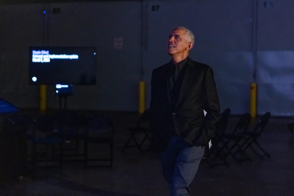 The image is a photograph of Barack Obama looking at the Power of Words prototype, which is not featured in the image. In the center Barack Obama stands, looking up at the photo and smiling. He is wearing a black blazer and denim blue jeans. His hands are in his pockets. Behind him to the left is a folding chairs in several rows and a monitor with indistinguishable words. The room is semi dark.