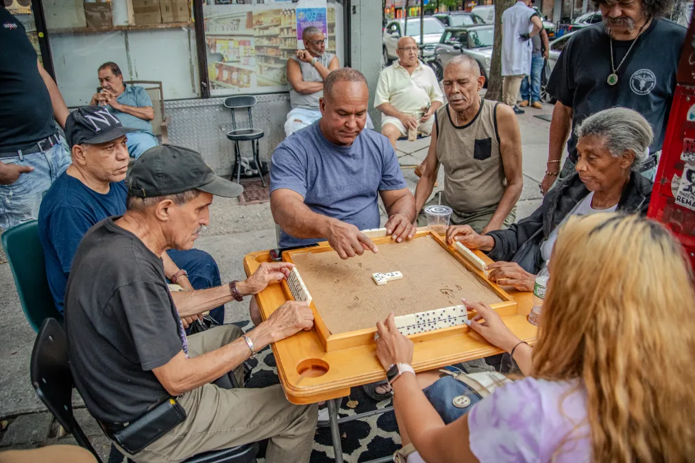 The image is a group of people in New York City sitting on the street, at a table, playing dominos. It is daylight outside. Behind the table is a storefront, a "bodega." At the table are four men and two women. All of the people at the table look to be elderly. From left to right: The first man has an olive complexion and is wearing a grey baseball hat and a grey shirt. He is holding onto to a row of dominoes in his hands. The second man has a olive complexion and is wearing a Yankees baseball cap and a blue shirt. He is staring at the table. The third man has a deep olive complexion and, a balding head with gray hair, and is wearing a blue shirt. He is pointing to the dominos table. The four man is sitting in a chair off to the side. He has an olive complexion, short grey hair, a grey goatee, and is wearing a sleeveless brown shirt with a black pocket on the left side of his chest. The fourth person is a women. She has a light deep complexion, long grey hair pulled back into a bun, and is wearing a blue denim jacket and a grey shirt. She is holding onto a row of dominos with both hands. The fifth person in the image is a woman. We cannot see her face, but we can see that she has long blond hair.