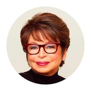 Valerie Jarrett has a soft, closed-lip smile. She has a light skin tone and short reddish brown hair. She is wearing red glasses and red lipstick.