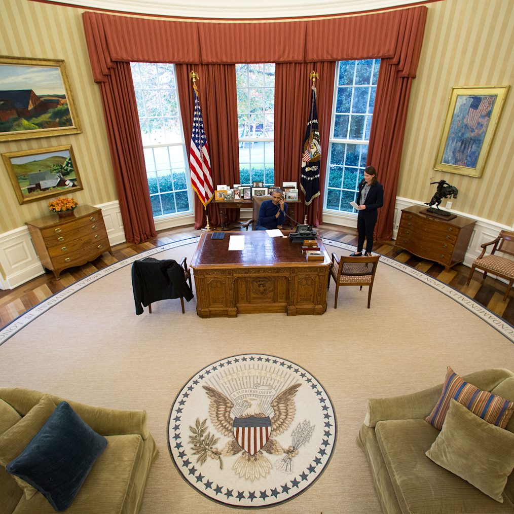 An aerial view of President Obama sitting at his desk in the Oval Office during his administration. There is a man standing to the right of President Obama. 