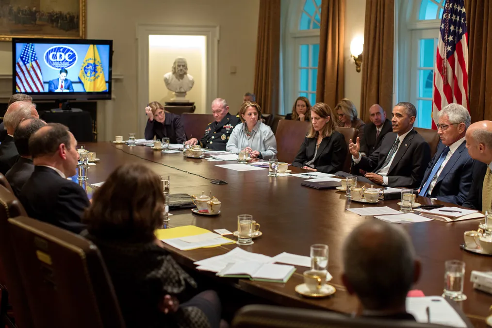 President Obama wearing a black suit and tie with a white button-up shirt sits at a long brown table with a group of individuals with a variety of skin tones. There is an American flag in the background on the right-hand side of the photo. There are two windows on a white wall with brown curtains. There is also a tv screening of someone sitting at a desk on the left-hand side of the photo.  