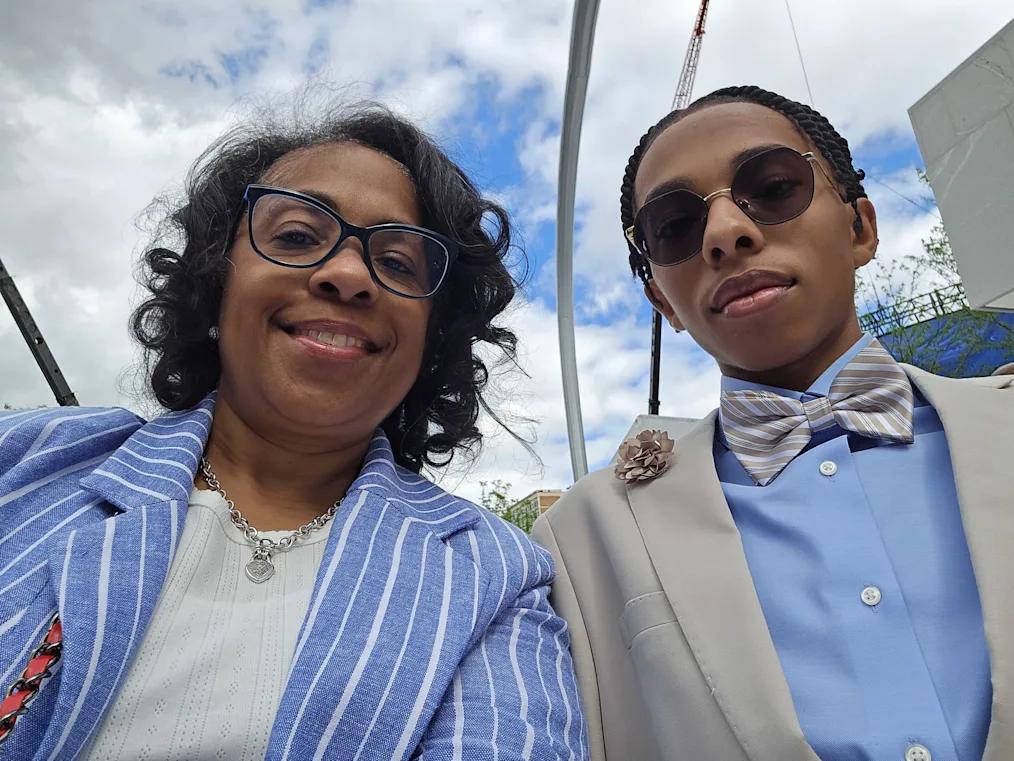 A low selfie of Michele and Jonathan Sullen. Michele is a Black woman with a medium skin tone and shoulder length curly black hair. Jonathan has a light skin tone and cornrows. The sky is bright with clouds. 