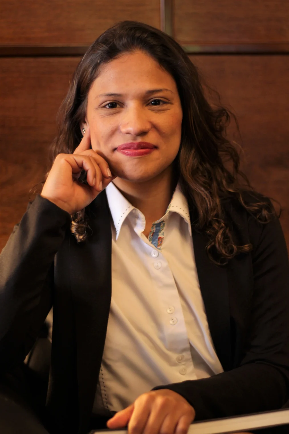 A young woman with long, wavy hair dressed in a white button-up shirt and black suit jacket poses for the camera. She has one hand on her cheek and one in her lap.