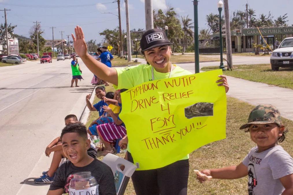 Sheila Babauta waves and smiles to camera, holding a neon yellow sign that reads "Typhoon Wutip Drive Relief." She is surrounded by smiling children of different skin tones.