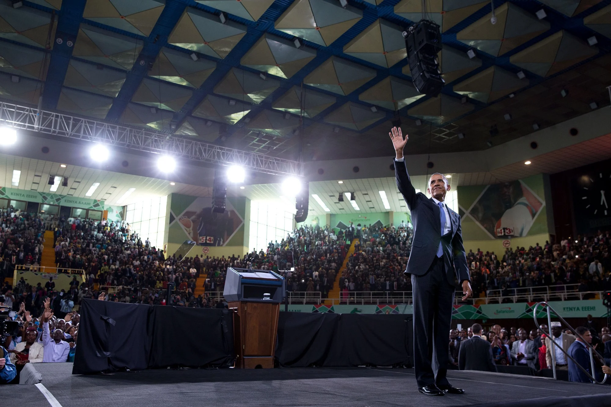 President Obama stands with his hand in the air facing a large crowd of people ranging from young to old and light to deep skin tones in the middle of a large room