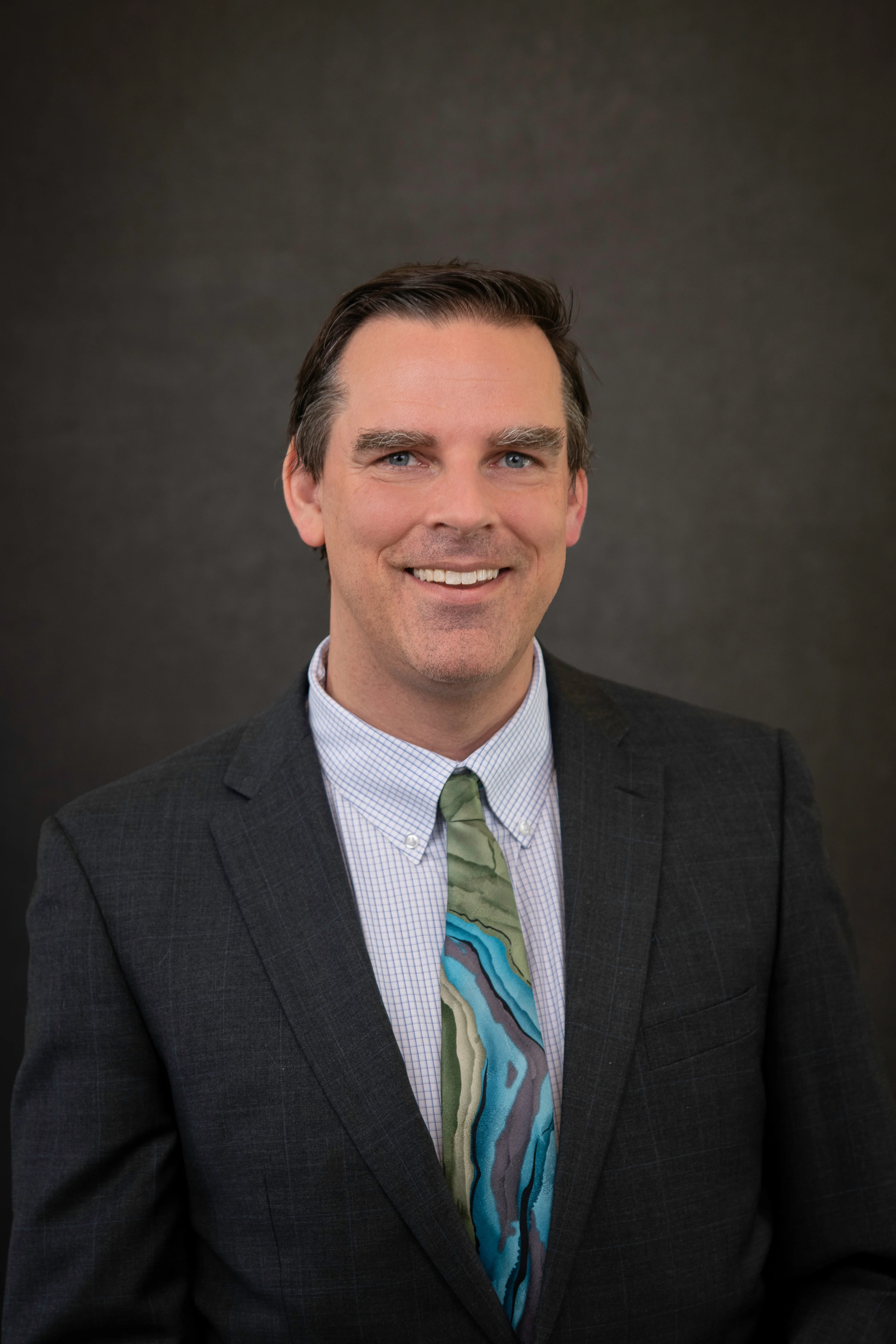 Jerad Morey, a man with a light skin tone and salt and pepper hair smiles at the camera. He is wearing a gray suit and a green and blue tie.