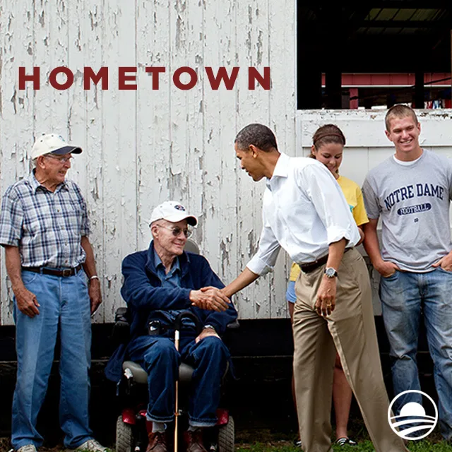 President Obama shakes a white man's hand. He has a with a light skin tone and is sitting along a wall with three others. The graphic reads, "HOMETOWN" at the top left corner in a dark red color.