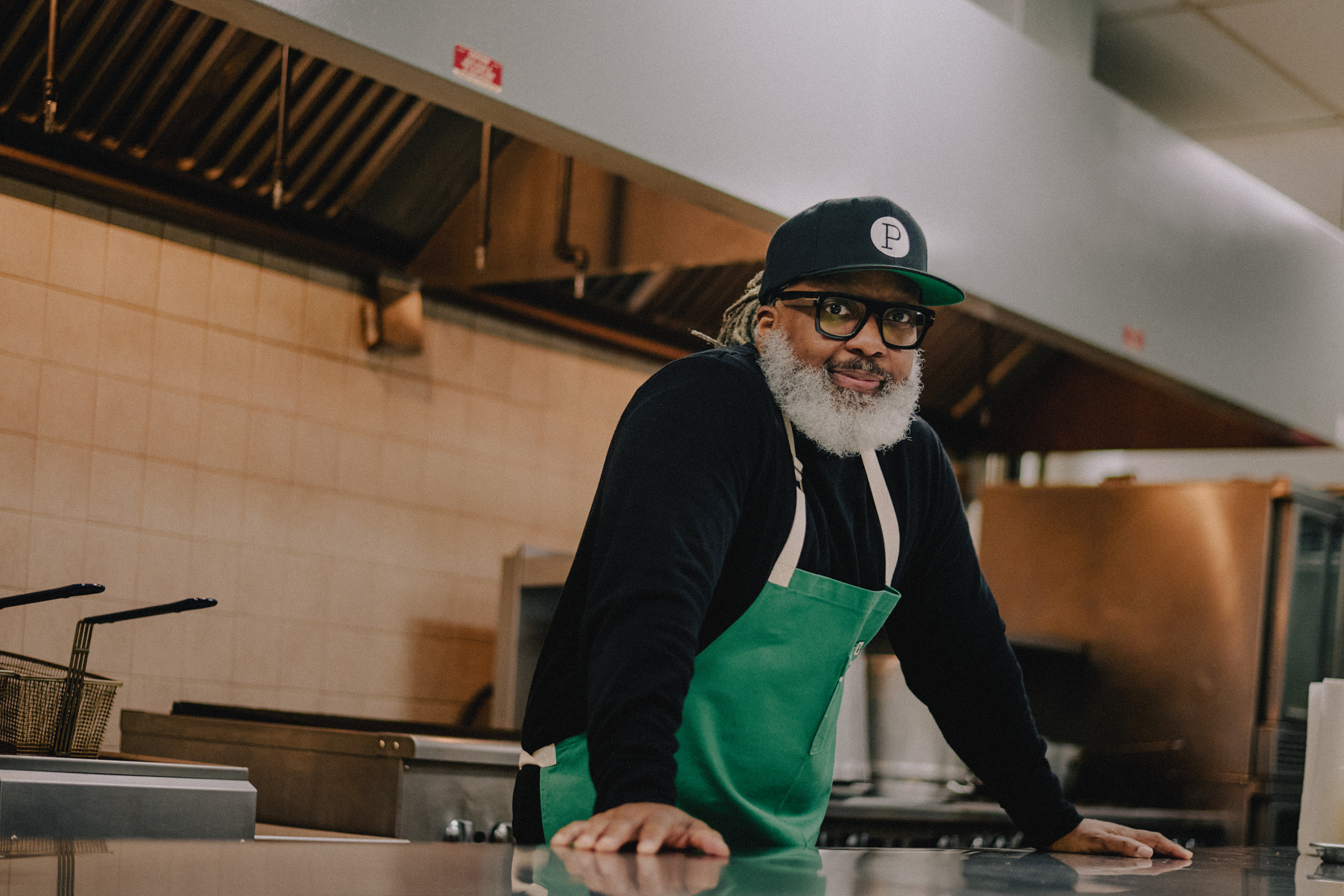 Chicago chef Cliff Rome, a Black man with a dark skin tone and long locs, looking directly at the camera. He has a gray beard. He is standing in a commercial kitchen. He is wearing a black baseball cap and a green apron. 