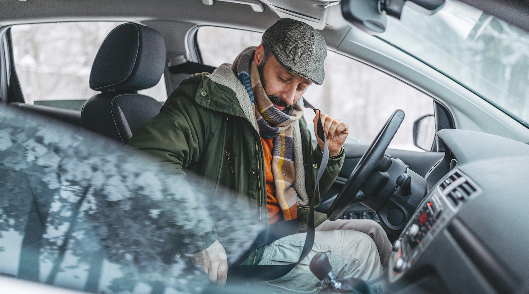 En person i grön jacka och grå keps sitter i förarsätet av en bil och spänner fast säkerhetsbältet.