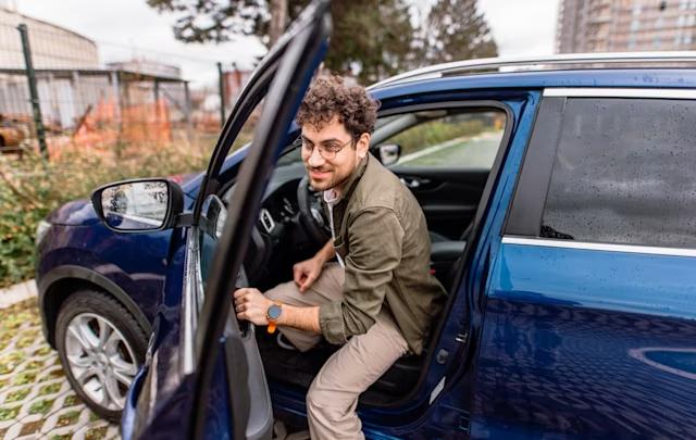 En man med glasögon och lockigt hår kliver ur en blå bil på en parkeringsplats.