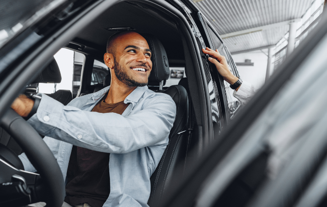 Person i ljusblå skjorta sitter i förarsätet på en bil och håller i dörren, fotograferad inifrån bilen.