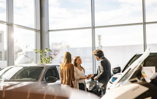 Tre personer samtalar i en ljus bilhall med stora fönster och solljus som strömmar in, omgivna av nya bilar.