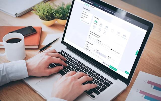 Person arbetar vid en bärbar dator med ett formulär på skärmen, bredvid ligger anteckningsbok, kaffekopp och papper med diagram.