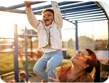 A woman holding up a small boy while he is hanging from the monkey bars at a park.