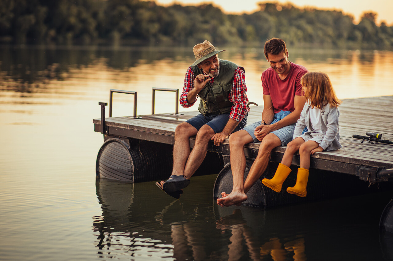 An old man, middle aged man and little girl sitting with their feet off the edge of a dock on a lake