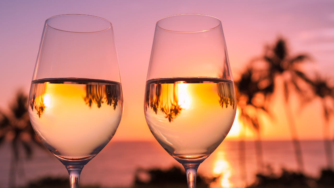 https:Two glasses of white wine clink together against a backdrop of a stunning sunset. The sky is ablaze with fiery oranges, pinks, and purples, and the silhouette of a palm tree sways gently in the breeze.