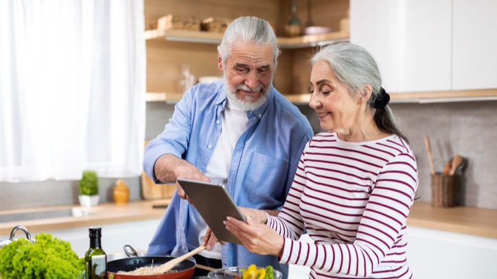 An elderly couple joyfully utilizing a digital tablet while in the kitchen.