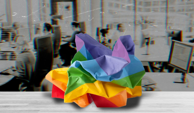 a crumpled rainbow-colored piece of paper in the foreground on an empty desk in an office, people at desks blurred in background