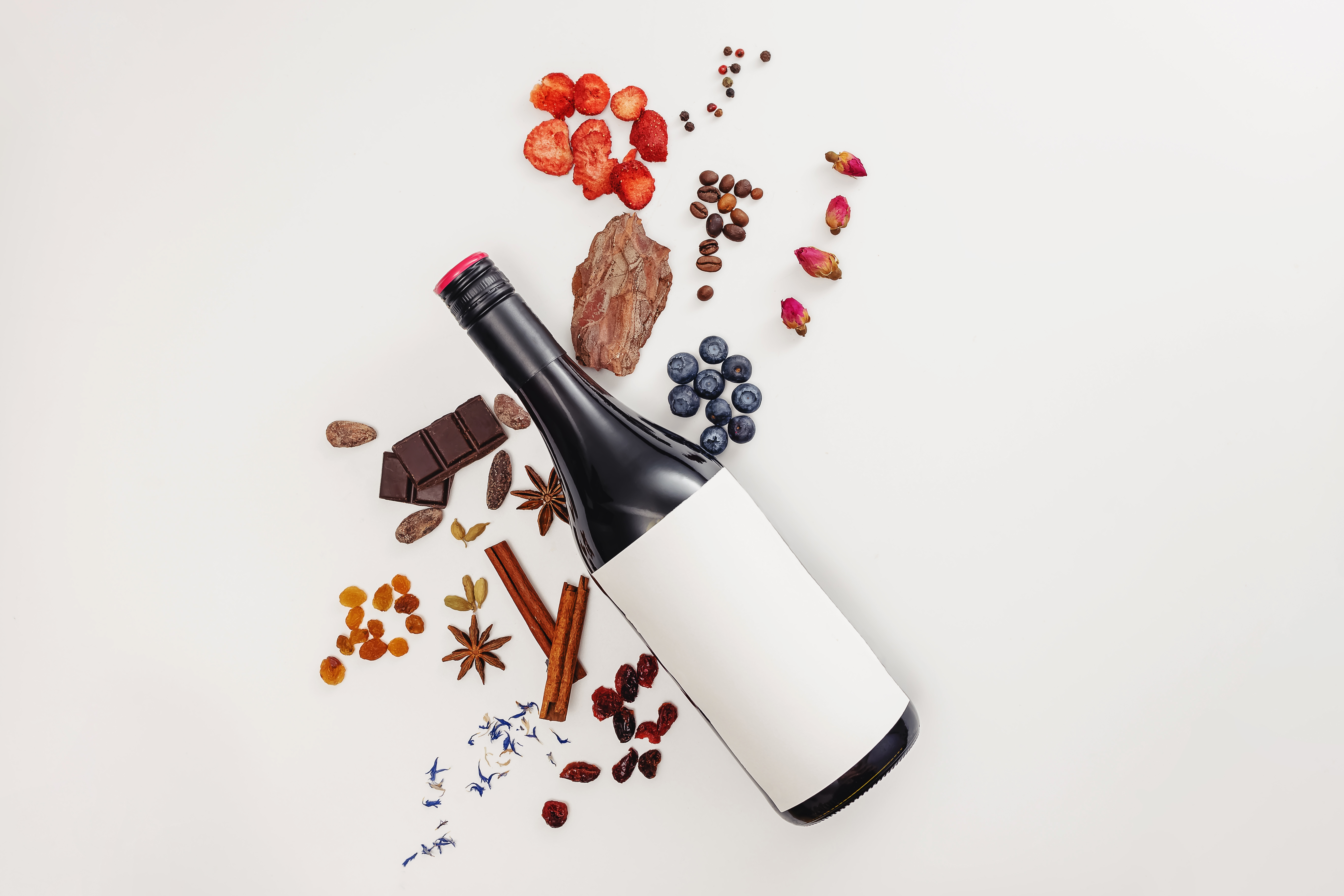 A bottle of red wine on a white surface with fruit, dark chocolate, raisins, and cinnamon sticks.