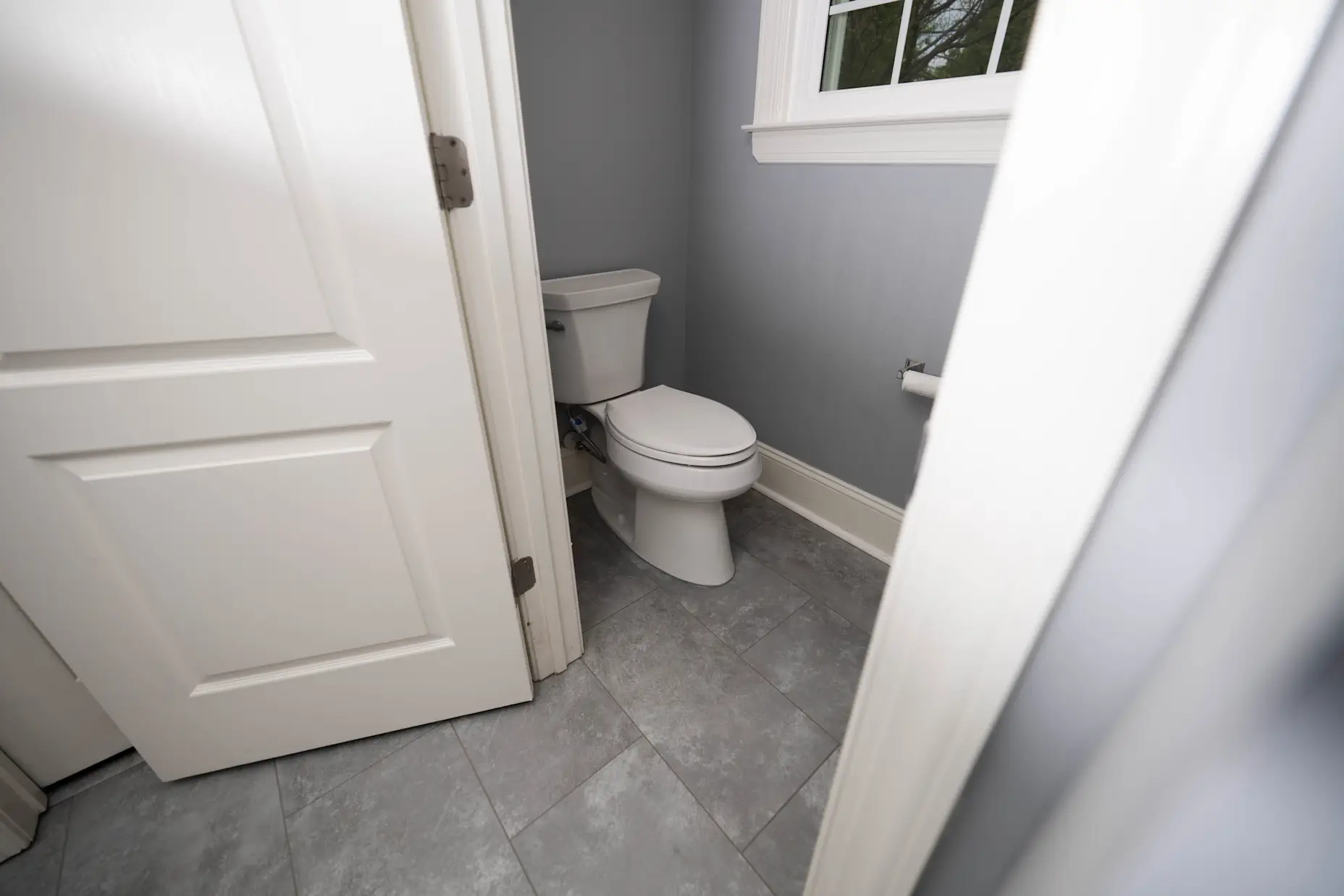 An image of an open door into a room with a white Kohler toilet and grey flooring.